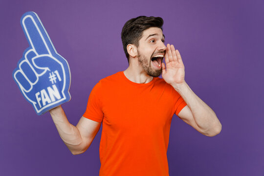 Young man he wear orange t-shirt cheer up support football sport team hold in hand fan foam glove finger up watch tv live stream scream aside with hands near mouth isolated on plain purple background