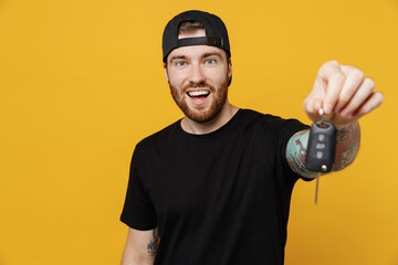 Young smiling happy cheerful cool bearded tattooed man 20s he wears casual black t-shirt cap hold in hand gives car keys fob keyless system isolated on plain yellow wall background studio portrait