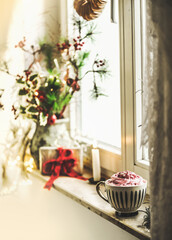 Cozy Christmas still life with cup of hot chocolate with whipped cream at window sill with seasonal winter decoration, gift and candle. Front view.