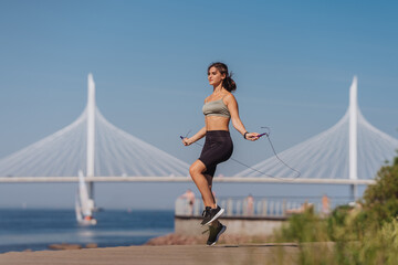 Girl jumps on a rope in a park against the background of a bridge across the bay. Morning workout of a Spanish girl on the beach. A young European woman does sports in nature on the weekend. Health.