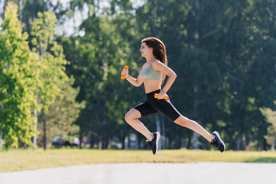 Smiley brunette hispanic young woman jumping high holding dumbbells against blurry park. Fit European girl training outside on summer sunny day, dressed in sportswear. Mock up of people at workout.