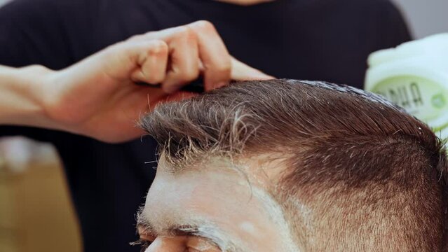 Barber applying hair gel to male client in barbershop close up