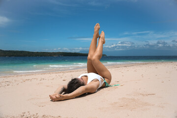 Fototapeta na wymiar A slim female in a white bikini is lying on the sand with her legs raised. Blue sky and sea in the background. Tourism in the Philippines. Happy vacation and free life style concept
