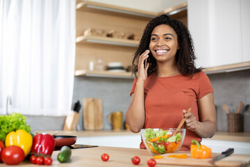Cheerful young african american woman preparing salad with organic vegetables speaks by smartphone