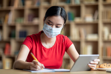 Serious busy young chinese girl in protective mask studying remotely with tablet in room interior
