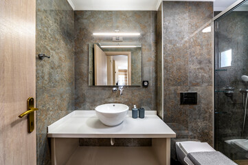 Modern bathroom in grey and brown colors with white bowl sink glass shower cabin in the corner, tap mounted in the wall, square mirror. Selective focus.