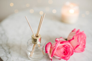 Obraz na płótnie Canvas Liquid scented home fragrance in glass bottle and fresh rose flowers over burning candle and glow lights in room. Cozy atmosphere.