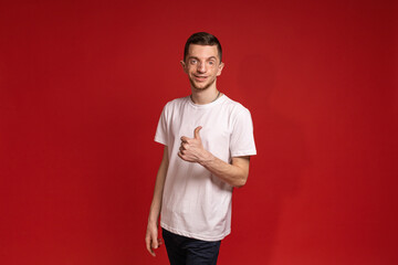 A young man in a white T-shirt stands on a red background and smiles while showing a thumbs up