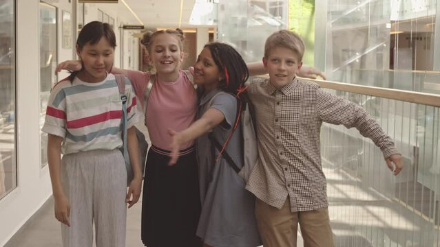 Medium slowmo of four cheerful diverse school children in casual clothes throwing paper airplanes at camera and hugging, standing in modern school corridor