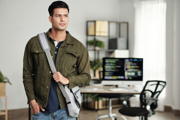 Portrait of young ambitious software developer with cross body bag standing in office and looking...