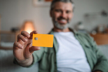 Smiling caucasian old man with beard shows credit card for finances in living room interior, selective focus