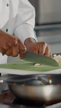 Vertical video: African american cook preparing garlic clove on cutting board, using organic aromatic ingredient to make gourmet dish in restaurant kitchen. Chef in uniform cooking professional meal