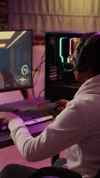 Over shoulder view of african american gamer using pc gaming setup relaxing playing first person shooter in multiplayer. Man streaming while explaining gameplay in online action game talking to team.