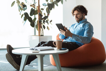Focused mature entrepreneur using digital tablet at workplace, sitting in bean bag chair at office, copy space