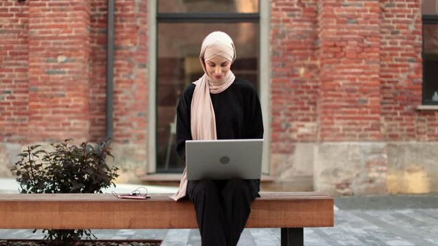 Cheerful young islamic businesswoman working on laptop while sitting near her office. Beautiful young woman with hijab working using laptop during break.