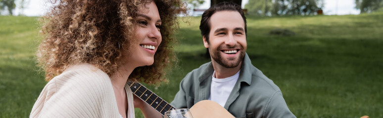 romantic man playing acoustic guitar near curly woman during picnic, banner.