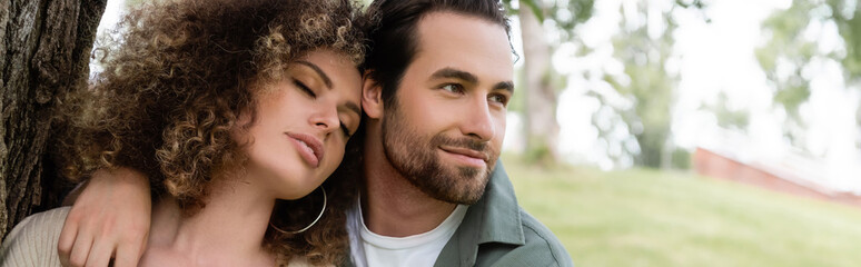 happy man embracing curly and young woman with closed eyes near tree trunk, banner.