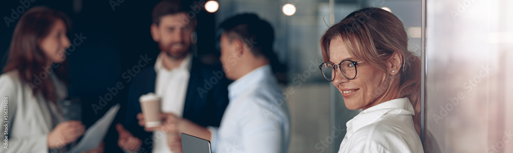 Wall mural Mature smiling business lady at office with group of colleagues on background, working on laptop.