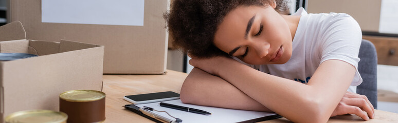 Obraz na płótnie Canvas tired african american volunteer sleeping near smartphone and clipboard in charity warehouse, banner.
