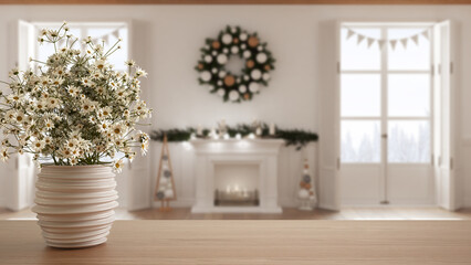 Fototapeta na wymiar Wooden table top or shelf with pottery vase with daisies, wild flowers, over Christmas living room with fireplace, windows on winter landscape, minimalist interior design concept
