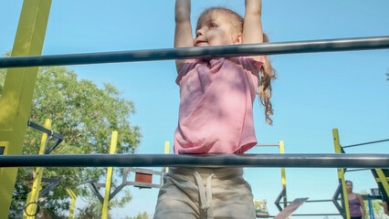 Obraz premium Little girl climbs gymnastic ladder on open sports ground on outside. Cute little girl crawls on vertical sports ladder in city park on sun day.