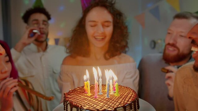 Close-up of delicious birthday cake and young woman in party hat blowing candles celebrating with noisy friends at home. Holidays and people concept.
