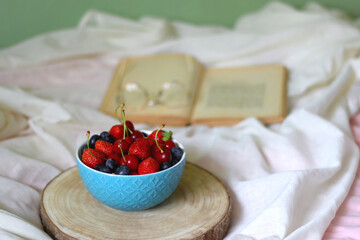 Bowl of strawberries, cherries and blueberries, open book and reading glasses on the bed. Selective focus.