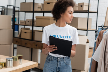 african american volunteer with clipboard looking at garments in charity center.