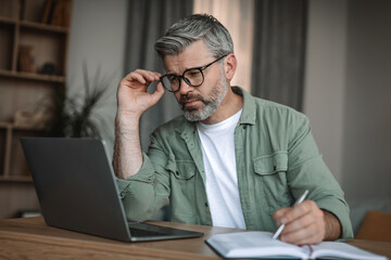 Busy serious european mature man teacher with beard in glasses looks at laptop, makes notes in room