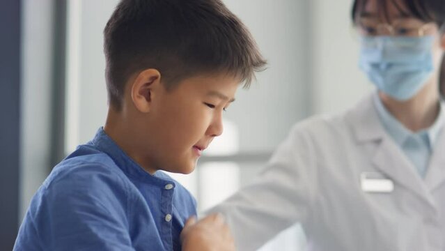 Sick Asian Boy Coughing As Female Doctor In Mask Taking His Body Temperature During Checkup In Medical Office