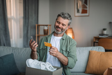 Glad caucasian old man with beard with cardboard box with sneakers uses credit card and smartphone