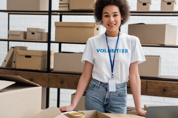 young african american volunteer with name tag smiling at camera near carton boxes.