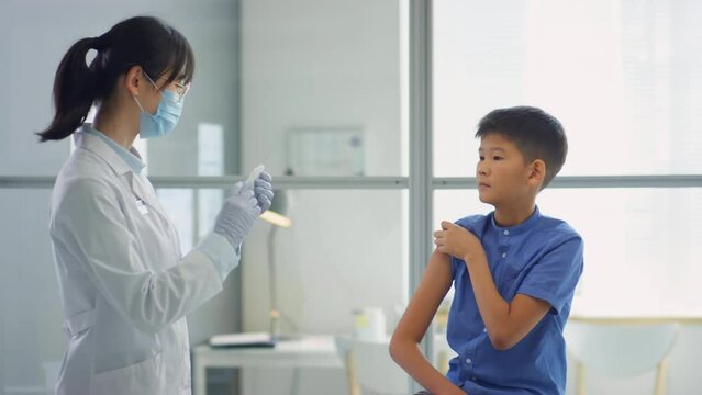 Female doctor in protective face mask and sterile gloves injecting anti-covid vaccine shot to Asian tween boy in clinic