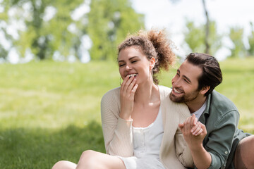 happy man and cheerful woman laughing while covering mouth in park.