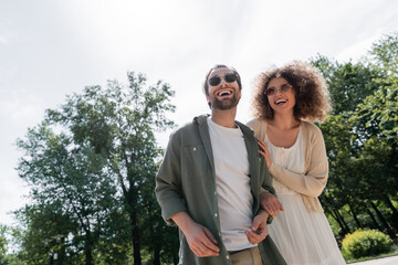 Fototapeta premium joyful man and happy curly woman in trendy sunglasses smiling in park.