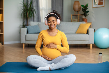 Happy young pregnant black female in earphones touching belly, relaxing on mat, practicing yoga and meditating