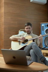 Cool guy playing acoustic guitar modern music looking at laptop, perform melody. Musical online lesson. E-learning