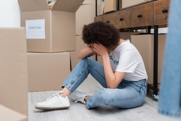 exhausted african american woman obscuring face with hands while sitting on floor in volunteer center.