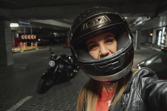 Young beautiful woman in a helmet takes a selfie in a parking garage in front of her motorcycle