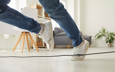 Clumsy person falls down after stumbling over a power cable. Man or woman in sneakers trips over an electrical cord while walking on the floor at home. Cropped shot. Domestic accident concept - obrazy, fototapety, plakaty