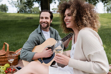 cheerful man playing acoustic guitar near happy woman with glass of wine during picnic.