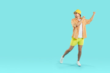 Cheerful carefree young man in summer clothes having fun on light blue background. Funny guy in shorts, shirt, panama and sunglasses dancing near copy space. Full length. Web banner.