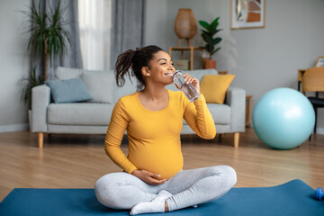 Cheerful pretty millennial pregnant african american lady practices yoga and drinks water from...