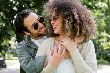 happy man in stylish sunglasses hugging pleased girlfriend in park.
