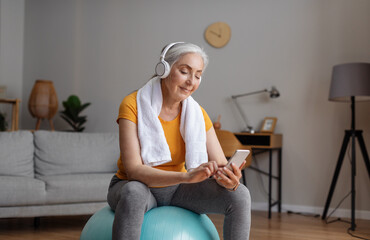Senior woman using smartphone and wearing headphones, sitting on fitball during online domestic workout