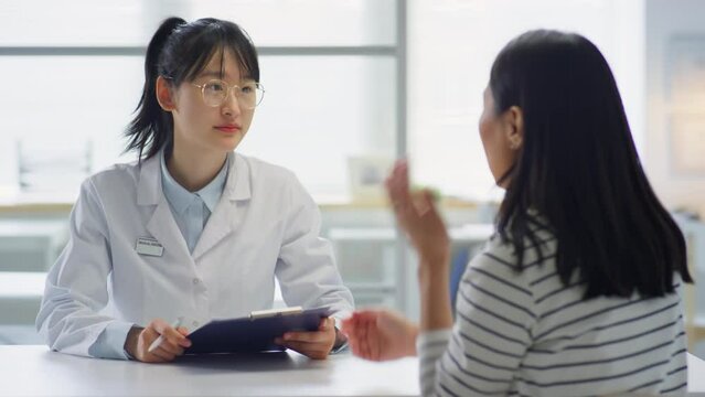 Asian female doctor in white coat holding medical history and giving consultation to woman while working in clinic