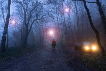 A man standing next to car watching glowing UFO's floating through trees. In a spooky forest on a foggy winters night.
