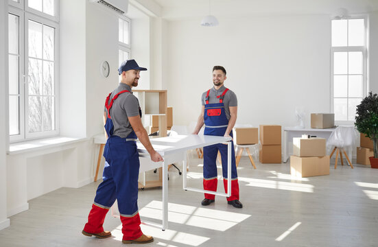 Loaders Carrying Furniture In Flat. Two Male Moving Service Workers Move Furniture When Moving To Office Or Apartment. Young Active Male Loaders In Blue And Red Overalls Carry White Desk.