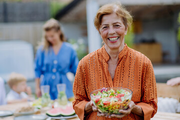 Happy senior woman serving salad at multi generation garden party in summer.