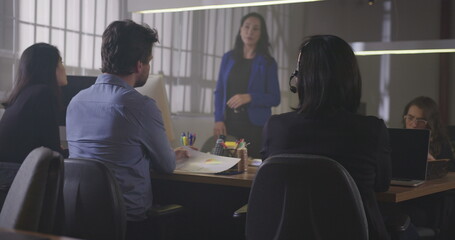 Group of young office workers gathered together for a meeting at night. Male employee sharing information with female co worker. Employees sitting at table in conversation
